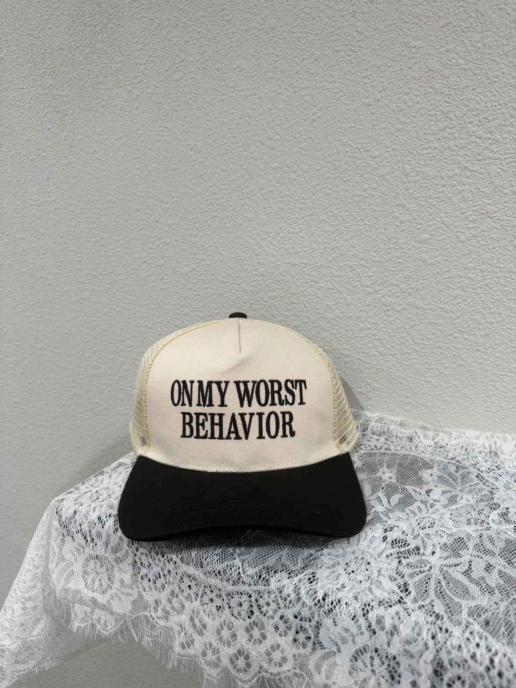A trucker-style cap with a cream front panel and black mesh back, printed with the text 'ON MY WORST BEHAVIOR' in black. The cap is styled on a white lace fabric against a textured light gray wall.