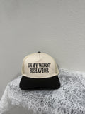 A trucker-style cap with a cream front panel and black mesh back, printed with the text 'ON MY WORST BEHAVIOR' in black. The cap is styled on a white lace fabric against a textured light gray wall.