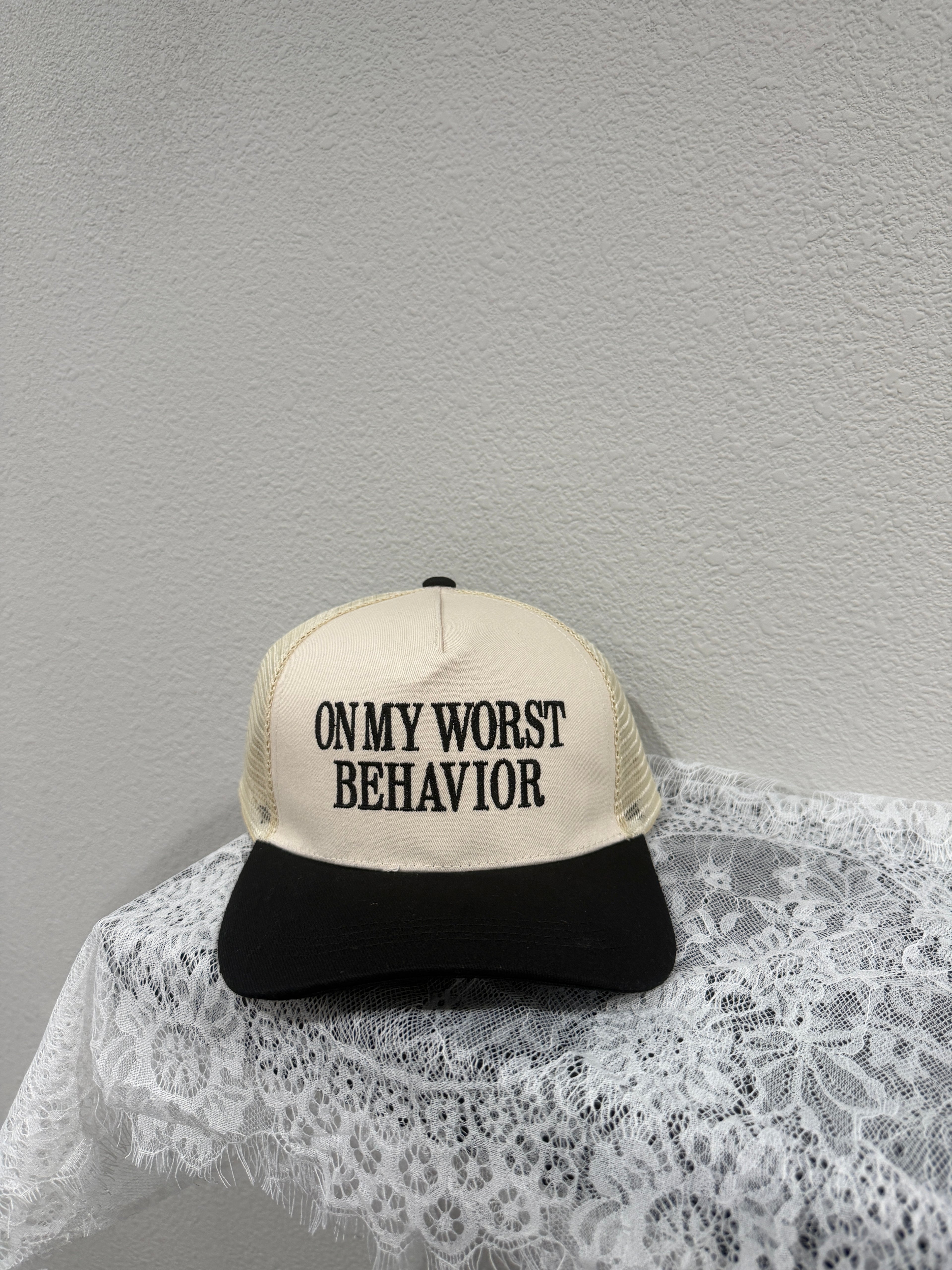 A trucker-style cap with a cream front panel and black mesh back, printed with the text 'ON MY WORST BEHAVIOR' in black. The cap is styled on a white lace fabric against a textured light gray wall.