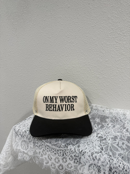 A trucker-style cap with a cream front panel and black mesh back, printed with the text 'ON MY WORST BEHAVIOR' in black. The cap is styled on a white lace fabric against a textured light gray wall.